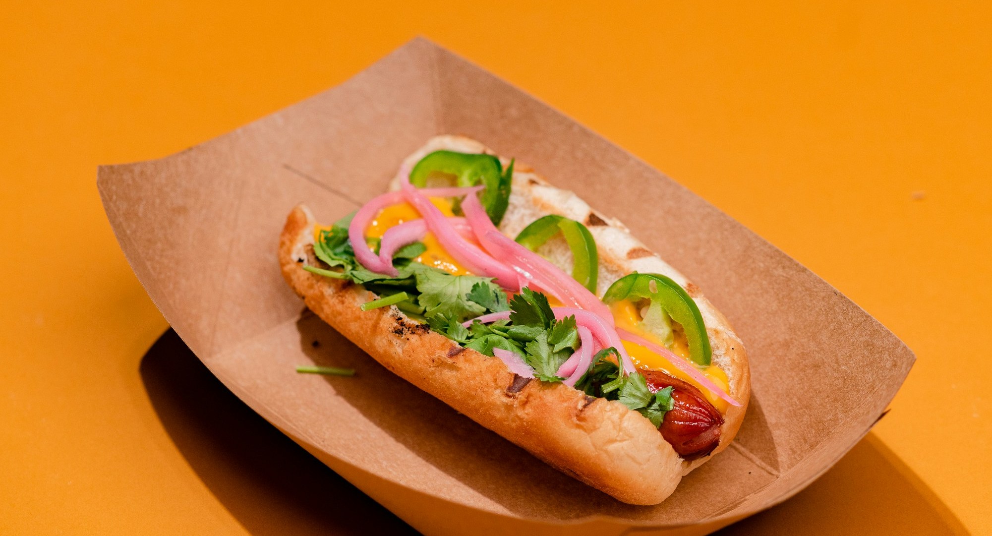 A gourmet hot dog with pickled onions, cilantro and jalapeños in a paper tray